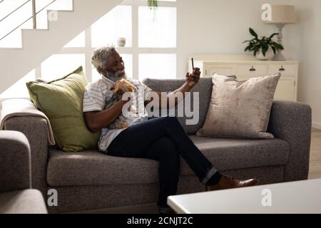 Afroamerikanischer älterer Mann, der auf einer Couch sitzt, ein Smartphone benutzt, ein Selfie macht, soziale Distanzierung und Selbstisolation in Quarantänesperre Stockfoto