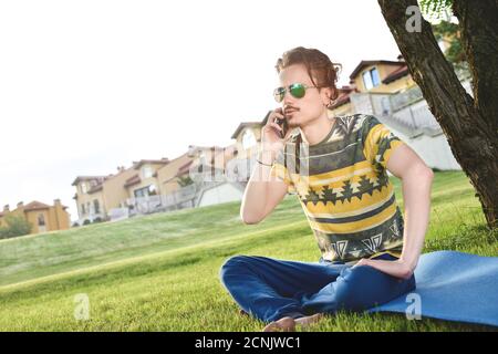 Junge stilvolle gutaussehende Mann sitzt auf Gras und sprechen auf dem Telefon ruhen im Park. Eine Pause einlegen Stockfoto