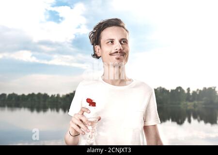 Junger Kerl trinkt frisches Wasser gegen den See und den Wald Hintergrund. Sommerdurst. Kerl will trinken Stockfoto