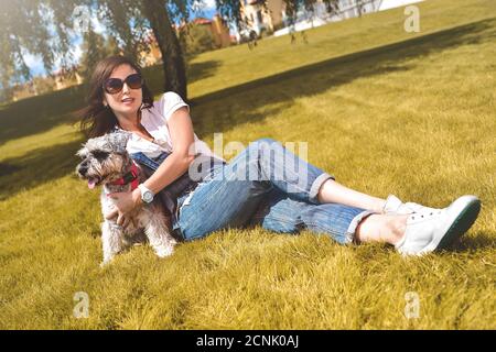 Hübsche Erwachsene kaukasische glückliche Frau, die sich an einem sonnigen Tag mit ihrem geliebten Hund im Park ausruhte. Weibchen mit Sonnenbrille liegt auf dem g Stockfoto