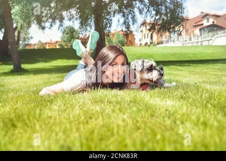 Hübsche Erwachsene kaukasische glückliche Frau, die sich an einem sonnigen Tag mit ihrem geliebten Hund im Park ausruhte. Weibchen liegt auf dem Gras, lächelnd an Stockfoto