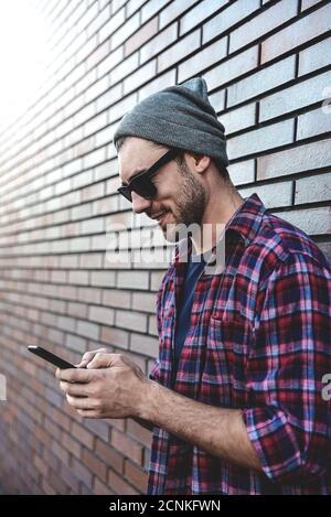 Textnachricht eingeben. Seitenansicht des hübschen jungen Mannes in eleganter Freizeitkleidung, der Mobiltelefon hält, während er sich an der Ziegelwand lehnt. Stockfoto