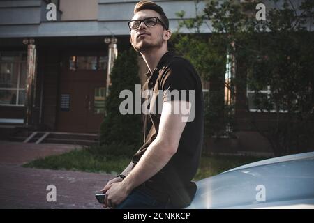 Junger Mann, der sich auf sein Auto stützt, ein Smartphone benutzt, lässig gekleidet. Stockfoto