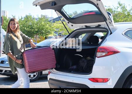 Junge glückliche Frau, die ihren Koffer in den Kofferraum des Autos packt. Urlaubskonzept. Stockfoto