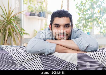 Lächelnder junger Mann posiert vor der Kamera, während er auf der sitzt Sofa Stockfoto