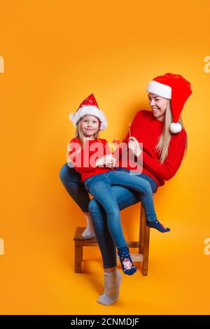 Fröhliche junge Mutter und nette Tochter Mädchen in Santa Hüte Halten Chistmas Süßigkeiten auf gelbem Hintergrund Stockfoto