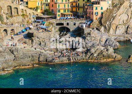 Schöne Nahaufnahme des Küstengebietes neben dem Yachthafen von Manarola. Die Menschen schwimmen in der Nähe der Felsformationen, wo das blau türkisfarbene Wasser... Stockfoto