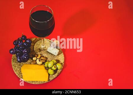 Nahaufnahme von einem Glas Rotwein, reifen Trauben, Nüssen, grünen Oliven und Käse auf rotem Hintergrund Stockfoto