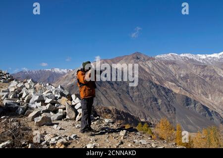 Fotograf und Model wandern an einem klaren Tag zu einem Hügel mit dem sichtbaren Mond, Nako Village, einem See, gelben Bäumen und strukturierten Bergen. Stockfoto