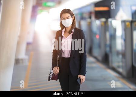 Asiatische Beifahrerin in einem schwarzen Anzug trägt medizinische Hygiene Schutzmaske allein stehend warten auf Zug am Bahnhof. Konzept für Geschäftsleute Stockfoto