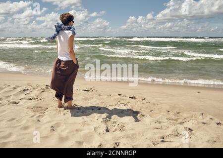 Frau mittleren Alters, die am Strand steht und das Winken anschaut Meer an windigen Sommertagen Stockfoto