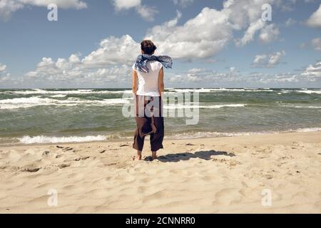 Einsam mittleren Alters stilvolle Frau von hinten stehend auf der Strand und Blick auf das Meer an windigen Sommertag Stockfoto