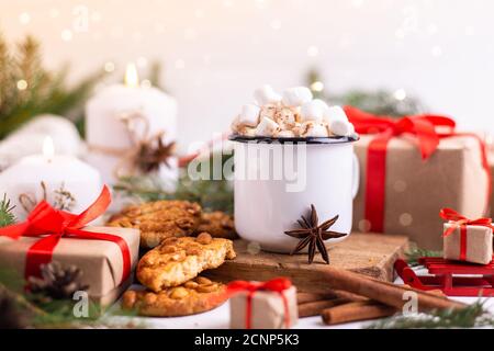 Emaillierte Tasse heißen Kakao oder Kaffee mit Marshmallows und Keksen. Rund um den Baum Äste, Geschenke und brennende Kerzen. Weihnachtsstimmung. Postkarte oder w Stockfoto