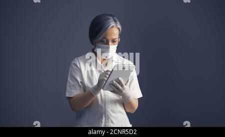 Maskierte Ärztin mit digitaler Tablette. Serious Grey haired Frau, die mit digitalen App oder Datenbank Stockfoto