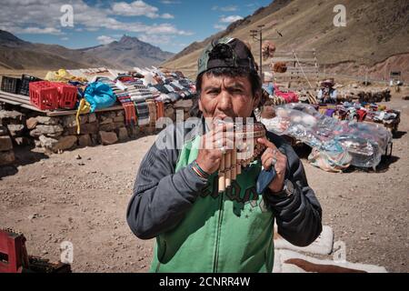 Ein Markthändler, der auf einem Straßenmarkt in den Anden von Altiplano, Peru, Panpipes Panpipes syrinx vorführt und Textilien und Stoffe verkauft Stockfoto