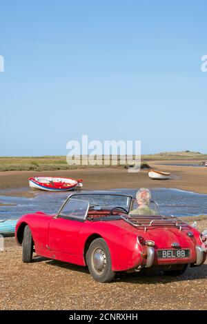 Boote festgemacht hoch und trocken bei Ebbe auf Sandbänken der Mündung. Mann sitzt in geparkten vintage Austin Healey. Klaren blauen Himmel Norfolk. Hochformat Stockfoto