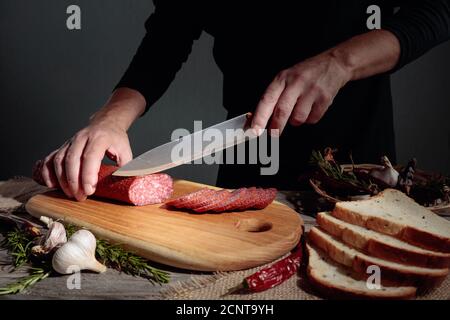 Mann schneidet eine Wurst für ein Sandwich. Geschnittene Salami mit Rosmarin, rotem Pfeffer und Knoblauch auf einem alten Holztisch. Stockfoto