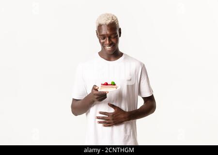 Portrait von zufrieden und glücklich afroamerikanischen jungen blonden Kerl, berühren Bauch zufrieden und Blick auf leckeren Kuchen, stehend weißen Hintergrund Stockfoto