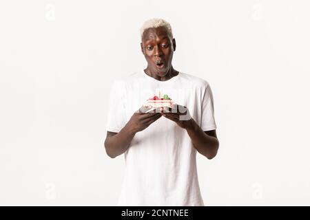 Porträt von gutaussehenden, fröhlichen afroamerikanischen blonden Mann, Blick amüsiert auf Stück Geburtstagskuchen, machen Wunsch und feiern b-day, weiß Stockfoto