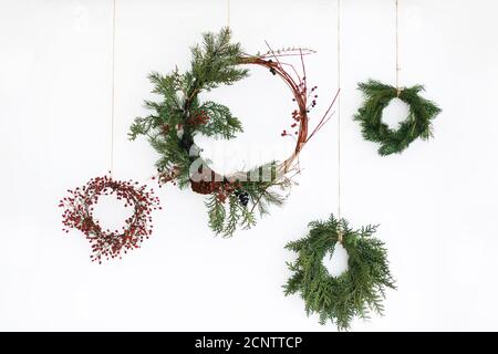 Rustikaler weihnachtskranz an weißer Wand, festliche Dekoration. Kreative natürliche und verschiedene weihnachtskränze mit roten Beeren und Tannenzweigen, Stockfoto