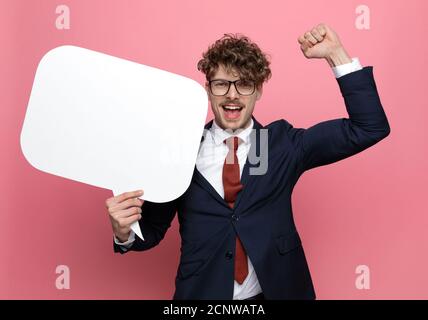 Fröhlicher junger Geschäftsmann in marineblauem Anzug mit Brille, Sprechblase haltend, Fäuste in der Luft haltend und den Sieg feiernden, lachenden und kreischenden Stockfoto