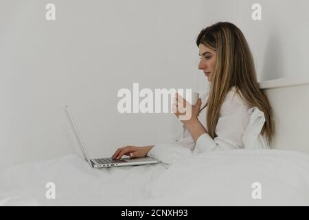 Junge schöne Frau mit einer Tasse Kaffee oder Tee mit Laptop-Computer auf dem Bett im Schlafzimmer entspannen. Arbeiten von zu Hause aus. Frau überprüft soziale App Stockfoto