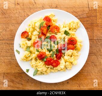 Ein Teller mit Rührei, gegrillten Tomaten und frisch geschnittenem Basilikum auf einem verzweifelten Holzhintergrund. Stockfoto