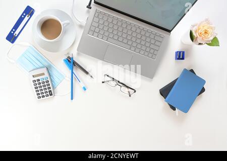 Weißer Schreibtisch mit silbernem Laptop, blauen Arbeitsmitteln und einer chirurgischen Gesichtsmaske gegen Coronavirus und Covid-19-Infektion, neues normales Geschäft Stockfoto