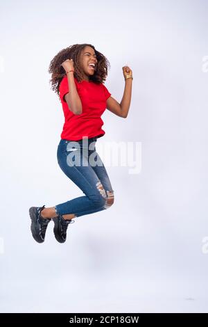 Hübsche junge afrikanische Frau trägt rote Top und blaue Jeans springen vor Aufregung und Glück, auf weißem Hintergrund Stockfoto