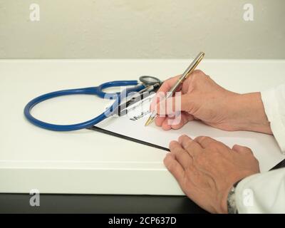 Hände des kaukasischen Arztes, der einen weißen Mantel trägt, der auf einer medizinischen Anamnese-Form mit einem Stethoskop auf dem Tisch schreibt. Stockfoto
