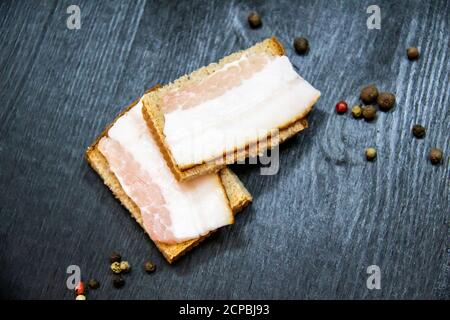 Auf dem Brot liegt ​​pieces Speck in Scheiben geschnitten. Traditionelles ukrainisches Essen. Auf dem Holzhintergrund liegen Scheiben gesalzener Schweinelardon auf dem Brot. Zwei l Stockfoto