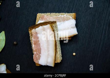 Auf dem Brot liegt ​​pieces Speck in Scheiben geschnitten. Traditionelles ukrainisches Essen. Auf dem Holzhintergrund liegen Scheiben gesalzener Schweinelardon auf dem Brot. Zwei l Stockfoto