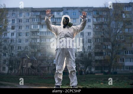 Zentrierte Momentaufnahme des Wissenschaftlers mit weißer Uniform, Handschuhen und Gasmaske, der vor einem hohen Wohnblock steht und mit beiden Händen Stoppschild zeigt, Konzept der gefährlichen natürlichen Umwelt Stockfoto