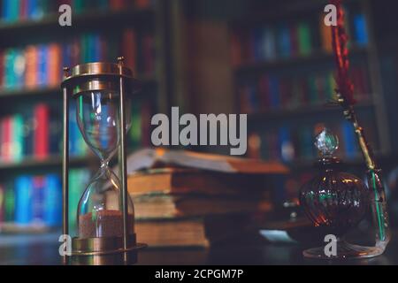 Hourglass und alter Federkiel, Sanduhr, Bücher und Vintage-Tintenfass auf Holztisch im alten Büro vor dem Hintergrund des Bücherregals. Konzeptionell Stockfoto