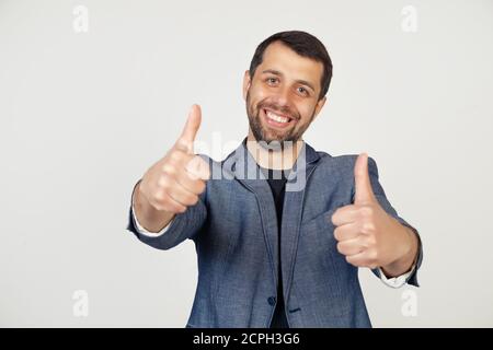 Junger Geschäftsmann mit einem Lächeln Mann mit einem Bart in einer Jacke, Genehmigung, macht eine positive Handbewegung, Daumen hoch lächelnd und glücklich für den Erfolg. Gewinnerbewegung Porträt eines Mannes auf grauem Hintergrund Stockfoto