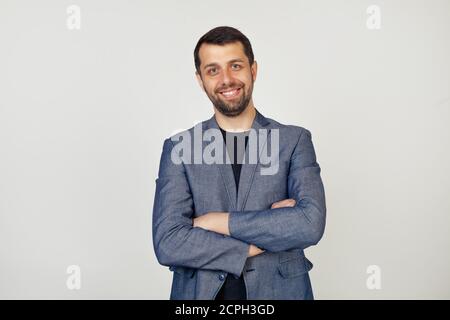 Junger Geschäftsmann mit einem Lächeln Mann mit einem Bart in einer Jacke, glückliches Gesicht, lächelnd mit gekreuzten Armen, Blick auf die Kamera. Eine positive Person. Porträt eines Mannes auf grauem Hintergrund. Stockfoto