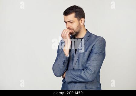Ein junger Geschäftsmann mit einem Lächeln, ein Mann mit einem Bart in einer Jacke, sieht angespannt und nervös mit den Händen auf den Lippen, beißt seine Nägel. Angstproblem. Porträt eines Mannes auf grauem Hintergrund. Stockfoto