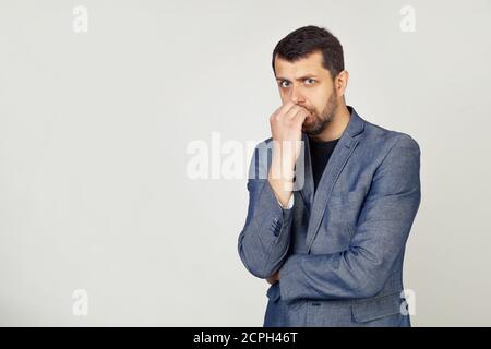 Ein junger Geschäftsmann mit einem Lächeln, ein Mann mit einem Bart in einer Jacke, sieht angespannt und nervös mit den Händen auf den Lippen, beißt seine Nägel. Angstproblem. Porträt eines Mannes auf grauem Hintergrund. Stockfoto