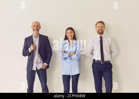 Drei fröhliche, mehrjährige Büroangestellte, die sich an die Wand lehnen und schauen Bei der Kamera Stockfoto