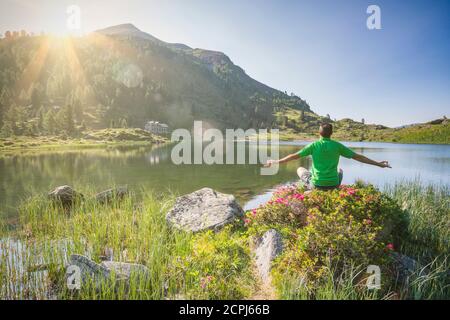 Mann 40 - 50 Jahre alt in entspannender Position bei sonnenaufgang am Ufer des Colbricon Alpensees in Sommer mit Rhododendron Blüte und Berg Stockfoto