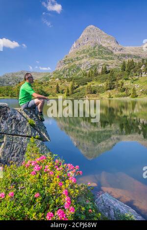 Colbricon Seen im Sommer mit Rhododendron Blüte und Berg reflektiert auf dem Wasser, ein Wanderer auf einem Felsen sitzen in einem Moment der Entspannung, Lagorai, Stockfoto
