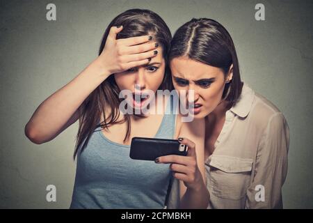 Zwei schockierte Frauen freuten junge Mädchen, die auf das Handy schauend Sehen schlechte Nachrichten oder Fotos mit ekelhaften Emotionen auf Gesicht isoliert auf grau wa Stockfoto