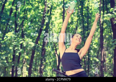 Gerne sorglos fit woman holding hands up mit einer Flasche Wasser in grün Sommer Park Stockfoto