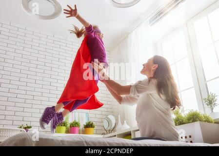 Mutter und ihr Kind spielen zusammen. Mädchen in einem Superhelden Kostüm. Das Kind hat Spaß und springt auf dem Bett. Stockfoto