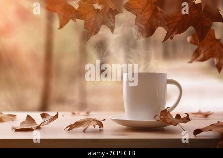 Tasse mit heißem Getränk auf einem Holztisch voll Trocken gefallene Blätter von Bäumen mit herbstlicher Landschaft Stockfoto