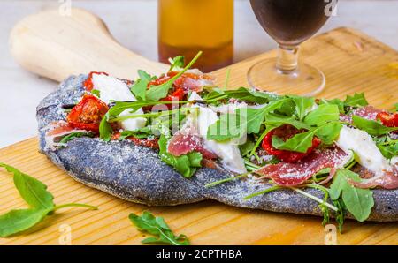 Schwarze Teig Pizza mit Schinken, Käse und Rucola Blätter auf einem Schneidebrett. Stockfoto