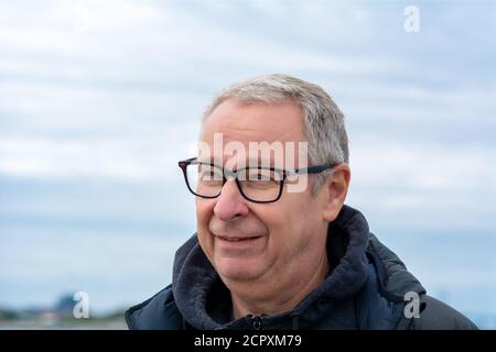 Nahaufnahme eines Mannes mittleren Alters, um die 50, mit grauen Haaren und Brille. Blauer Dunsthimmel im Hintergrund Stockfoto