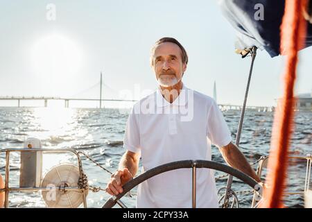 Reifer Mann steuert eine private Yacht während im Urlaub. Bärtiger Segler, der auf seinem Segelboot steht und in die Ferne blickt. Stockfoto