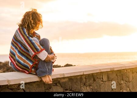 Unkenntlich einsame Frau setzen sich auf eine Wand suchen und Genießen Sie den Sonnenuntergang am Meer - farbige trendige mexikanische Poncho-Stil und Menschen im Freien Stockfoto