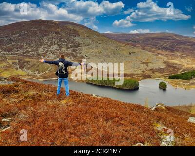 Ein Mann, der die Hände aufhebt und die Arme öffnet. Erkunden Sie das Hochland Mountain, Mann mit Blick auf einen See im schottischen Hochland. Stockfoto